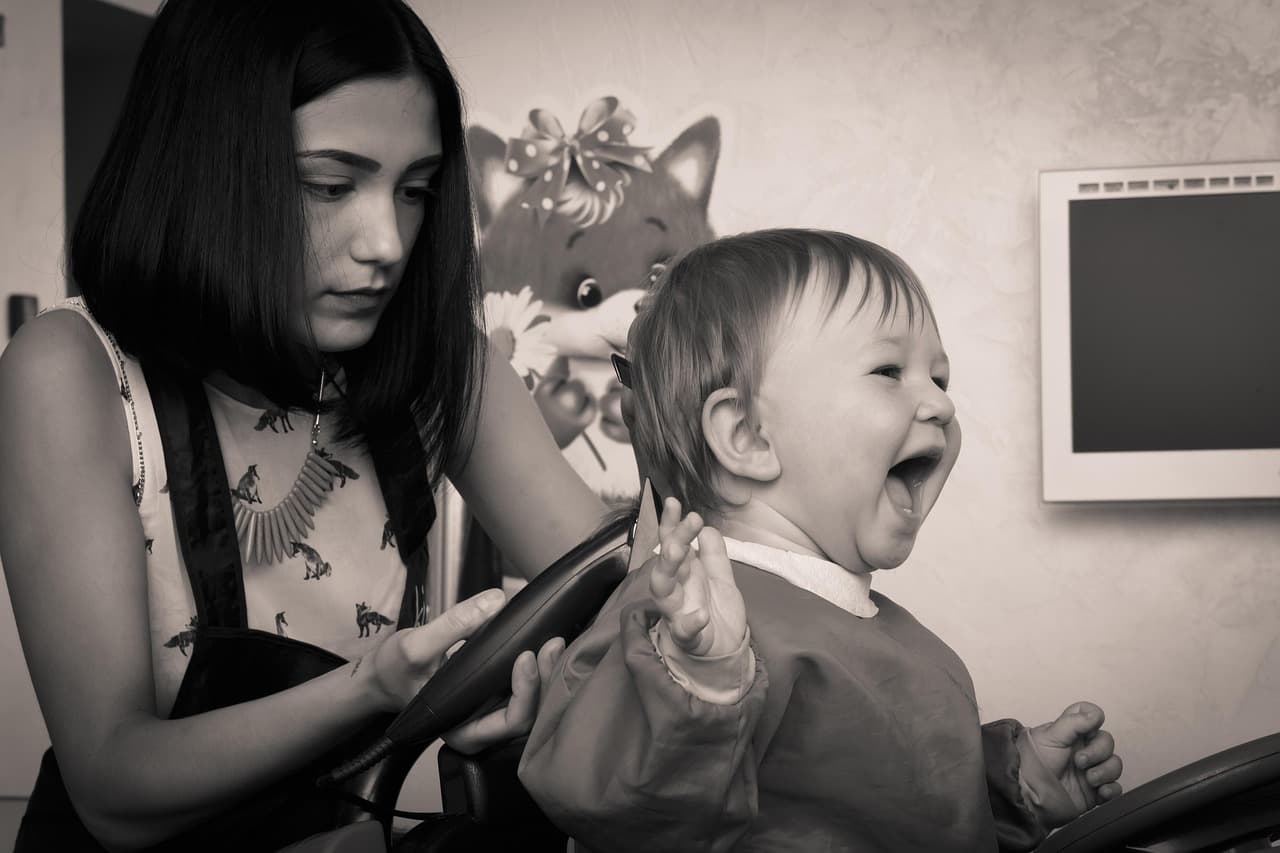A toddler laughing in a toy-car chair as the hairdresser starts his haircut