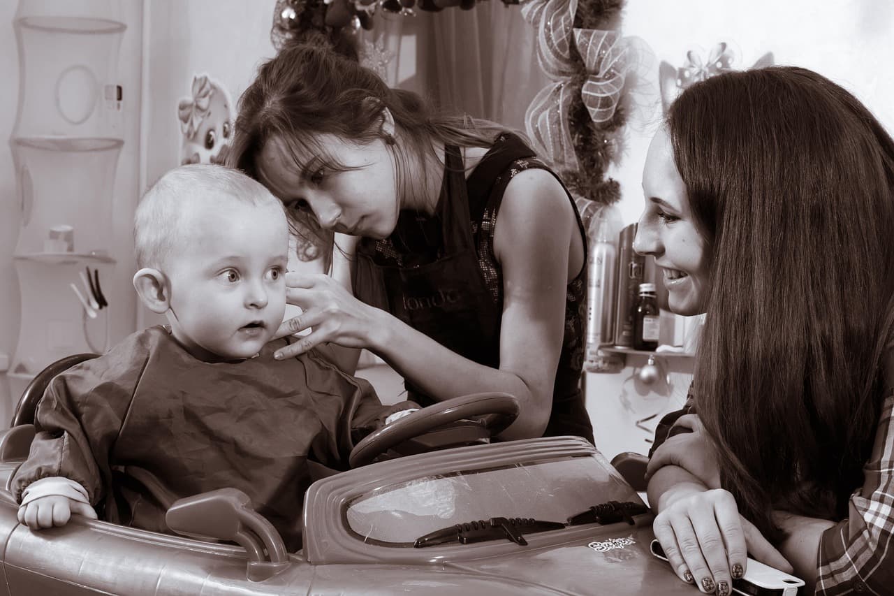 A mum smiling at her baby while a hairdresser trims round his ear