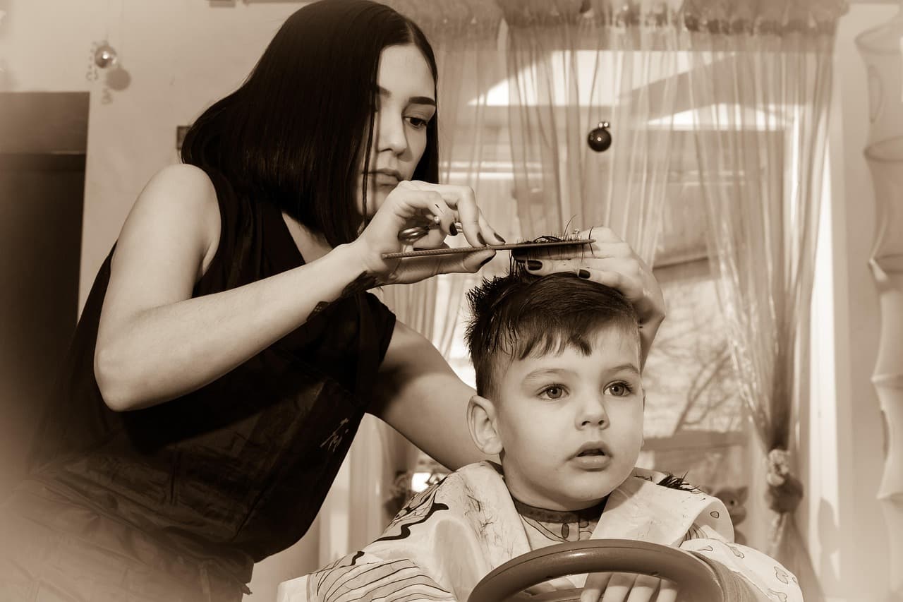 A hairdresser trimming a young boy's hair with comb and scissors while he sits in a toy-car chair